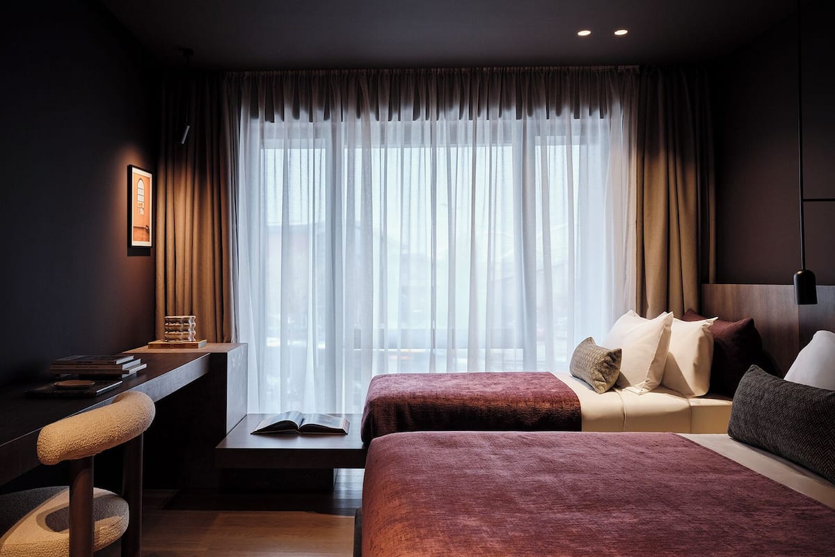 Hotellihuone, jossa on suuri ikkuna, työpöytä tuolilla, kaksi sänkyä ja yöpöytä.