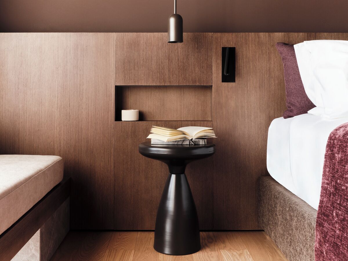 A compact hotel room with a bed, a small black table with an open book, a white pillow, and a wooden wall.