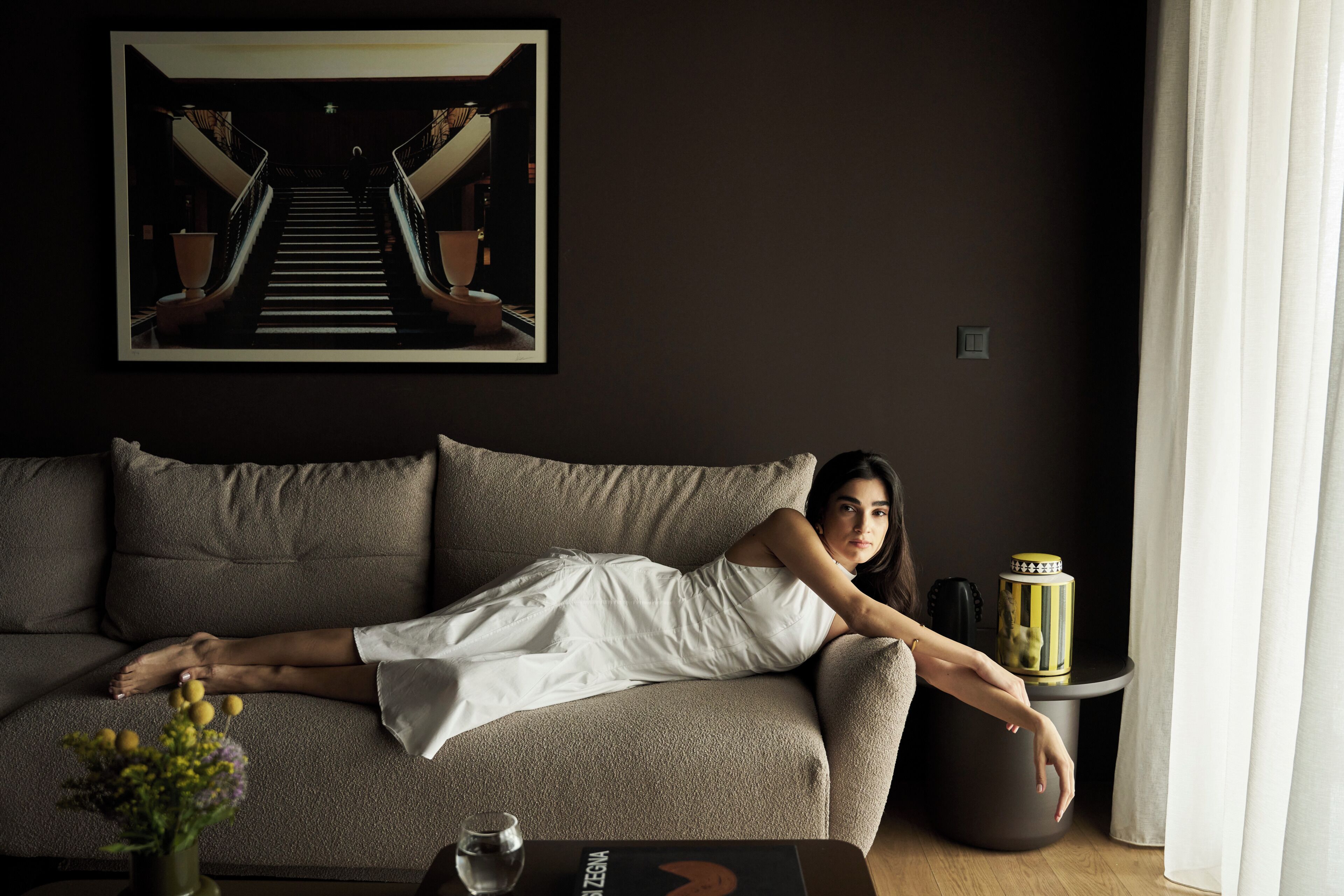 A person lying on a sofa in a room with a framed picture on the wall, a small table with a vase and a glass, and a round side table with a decorative item.