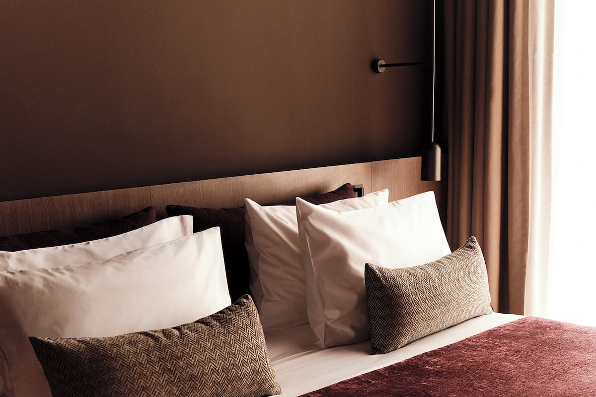 Hotellihuone, jossa on puinen sängynpääty, valkoisilla ja ruskeilla tyynyillä varustettu sänky sekä punainen heittopeitto.