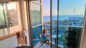 Ein Balkon mit Meerblick, ein Kind sitzt an einem Tisch, und ein Stuhl steht daneben.