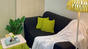Una habitación pequeña y acogedora con un sofá negro, una manta blanca y una planta verde. Hay una mesita con un libro y una lámpara. Las paredes están decoradas con obras de arte sencillas.