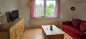 A living room with a wooden cabinet, a red sofa, a coffee table, a TV, and a window with checkered curtains. - Apartment 'Wiesmoor - Ranch' with Private Terrace, Shared Garden and Wi-Fi (Wiesmoor)