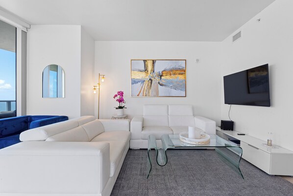 Una sala moderna con un sofá blanco, un sofá azul, una mesa de centro de vidrio y un televisor de pantalla plana montado en la pared.