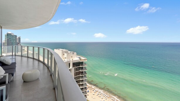 A balcony with a view of the ocean and beach, furnished with a sofa, ottoman, and chairs.