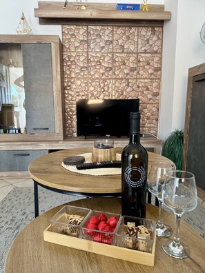 A modern living room with a stone fireplace, a television, a round wooden table with snacks, wine glasses, and a bottle of wine.