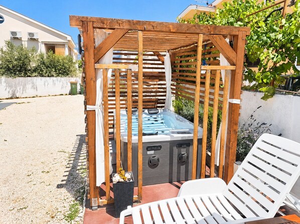 A wooden pergola structure with a hot tub underneath, surrounded by a gravel area and a white lounge chair.