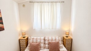 Un dormitorio con una cama, dos mesitas de noche, un ventanal con cortinas y dos lámparas.