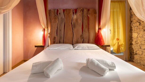 A large bed with white linens and two towels, a wooden headboard, red and yellow curtains, and a stone wall.