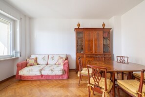 A room with a wooden dining table, four chairs, a sofa with floral cushions, and a wooden cabinet with glass doors. - Porto Vecchio Apartment by Wonderful Italy (Sanremo)
