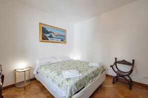 A bedroom with a bed, a bedside table, a chair, a painting, and a lamp. - Porto Vecchio Apartment by Wonderful Italy (Sanremo)