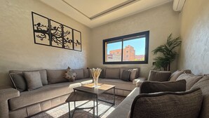 A modern living room with a sectional sofa, a coffee table, and a large window providing a view of buildings. - Charming 2-bedroom apartment in lovely Marrakesh with AC, Pool (Marrakesh)