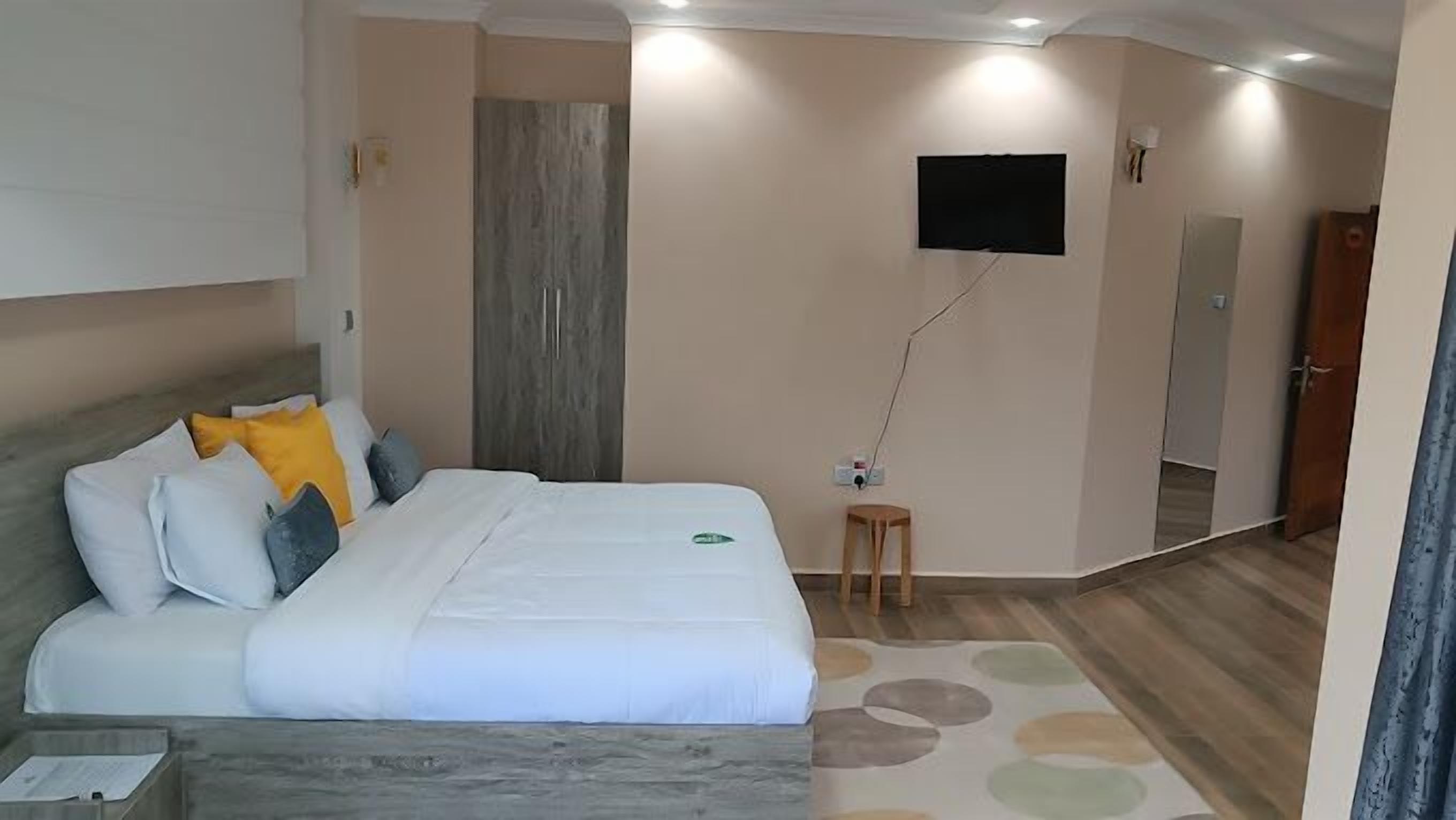 A hotel room with a bed, a small wooden stool, a flat-screen TV mounted on the wall, and a mirror.