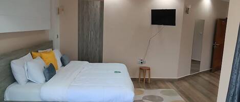A hotel room with a bed, a small wooden stool, a flat-screen TV mounted on the wall, and a mirror.