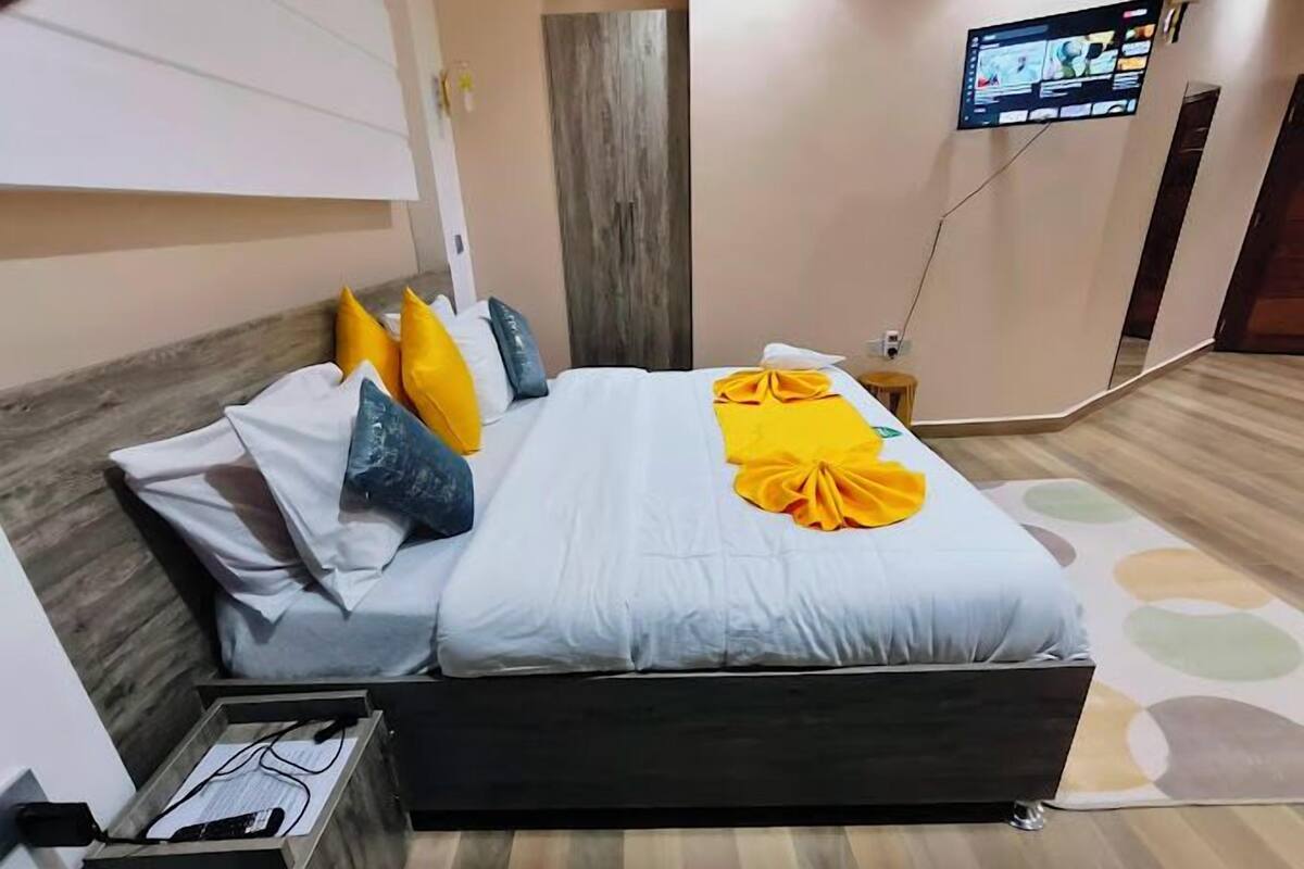 Une chambre d’hôtel moderne avec une tête de lit en bois, un téléviseur à écran plat fixé au mur et un lit recouvert d’une couette blanche et agrémenté de coussins colorés.