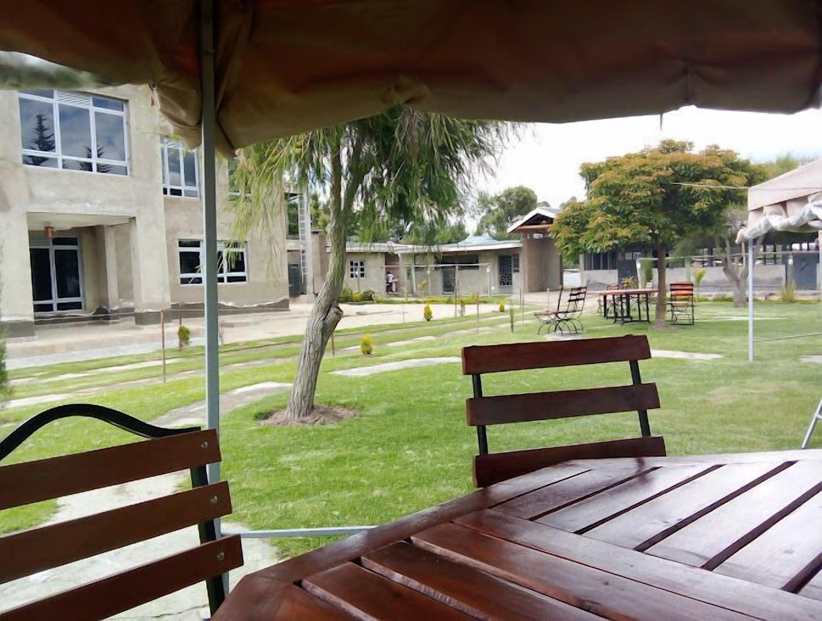 Uma área externa com mesas e bancos de madeira, cercada por um gramado bem cuidado e edifícios ao fundo.