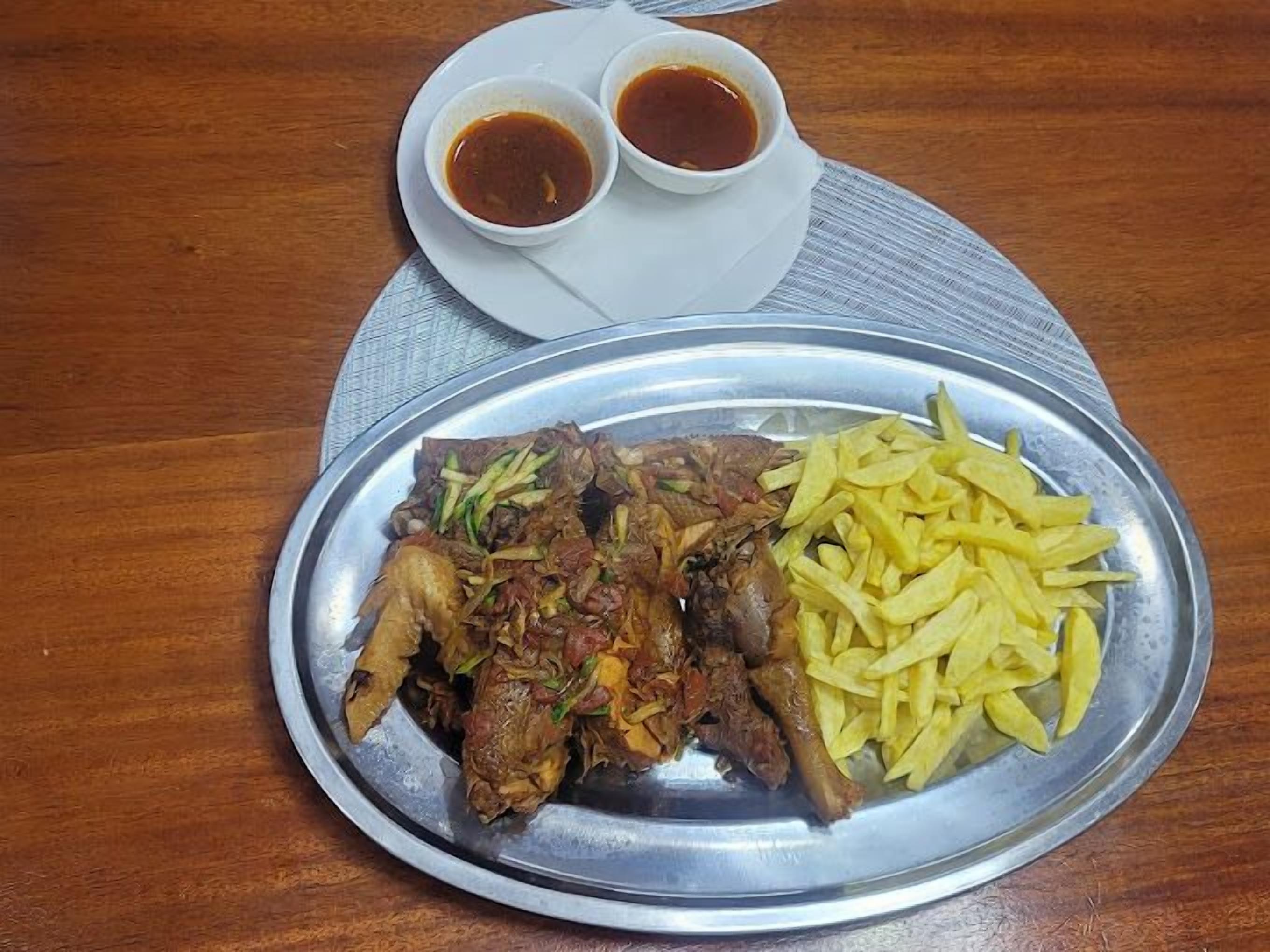 A meal consisting of grilled meat, fries, and two small bowls of sauce on a metal plate, served on a wooden table.