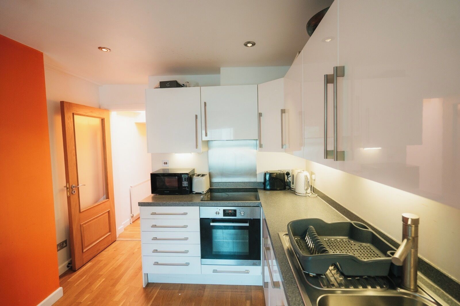A compact kitchen with white cabinets, a built-in oven, a microwave, a toaster, and a sink with a kettle.