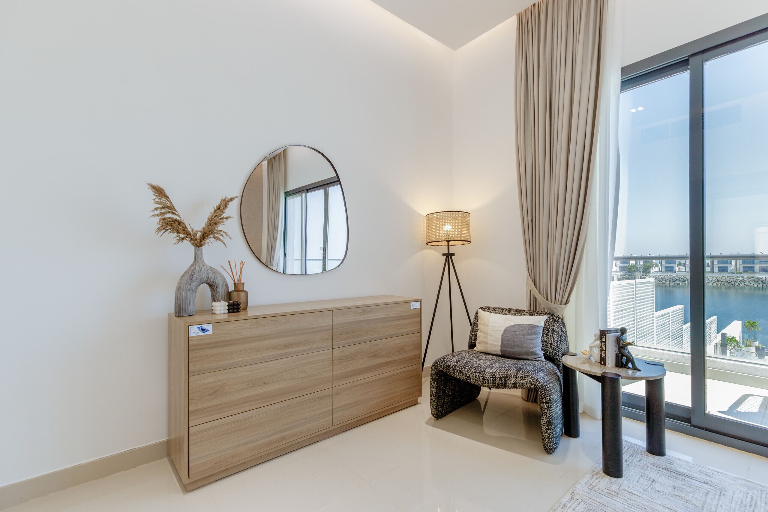 Ein modernes Wohnzimmer mit einem Holzkasten, einem runden Spiegel, einer Stehlampe, einem gemusterten Sofa und Blick auf einen Hafen durch große Fenster.