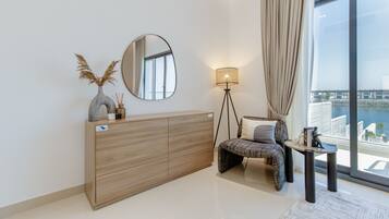 Ein modernes Wohnzimmer mit einem Holzkasten, einem runden Spiegel, einer Stehlampe, einem gemusterten Sofa und Blick auf einen Hafen durch große Fenster.