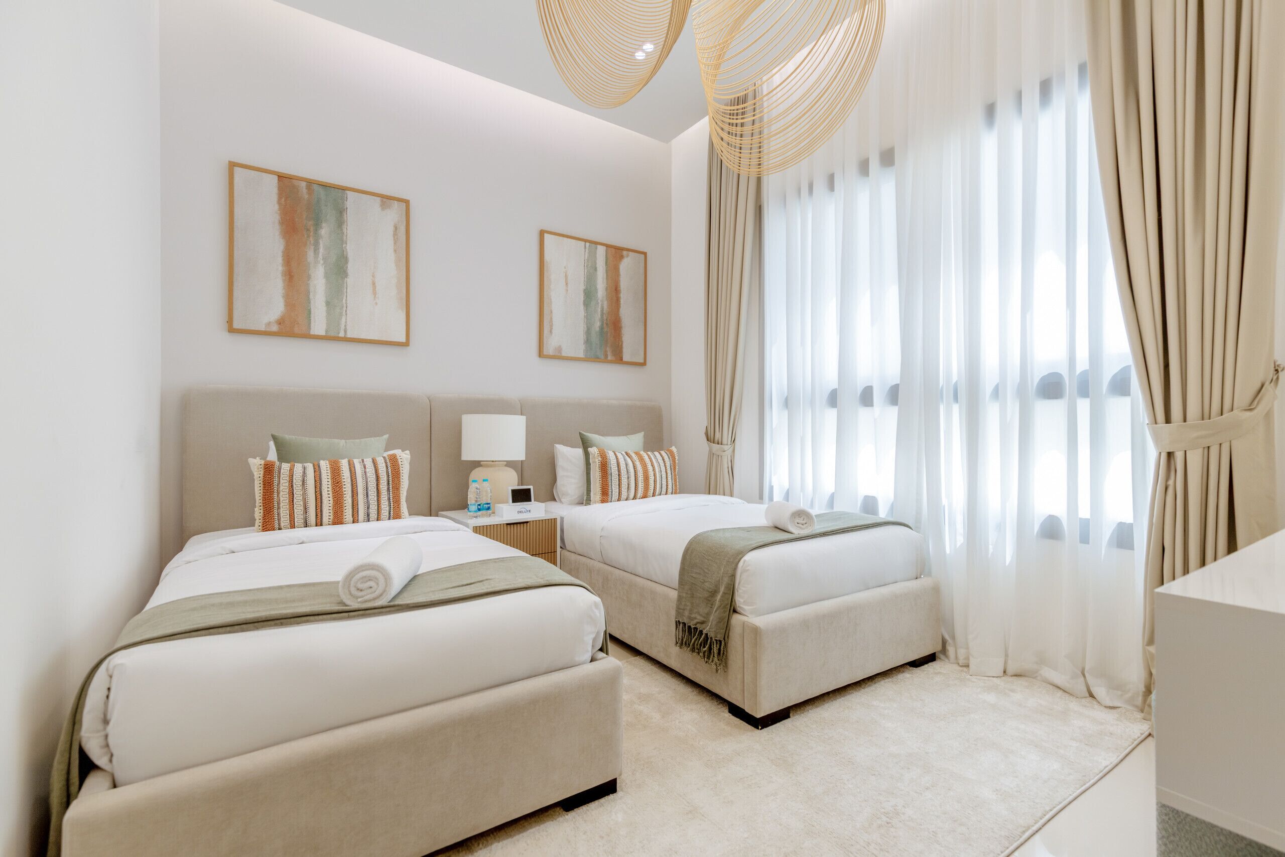 Une chambre d’hôtel avec deux lits, une table de chevet avec une lampe et une grande fenêtre avec des rideaux légers.