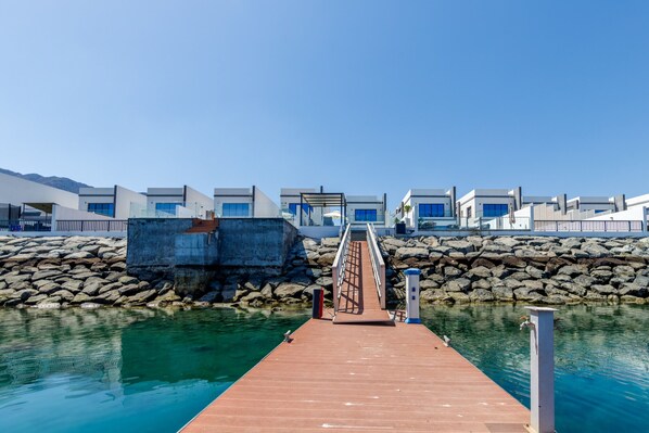 A wooden pier leading to a modern waterfront hotel with a clear view of the sea and a rocky shoreline.