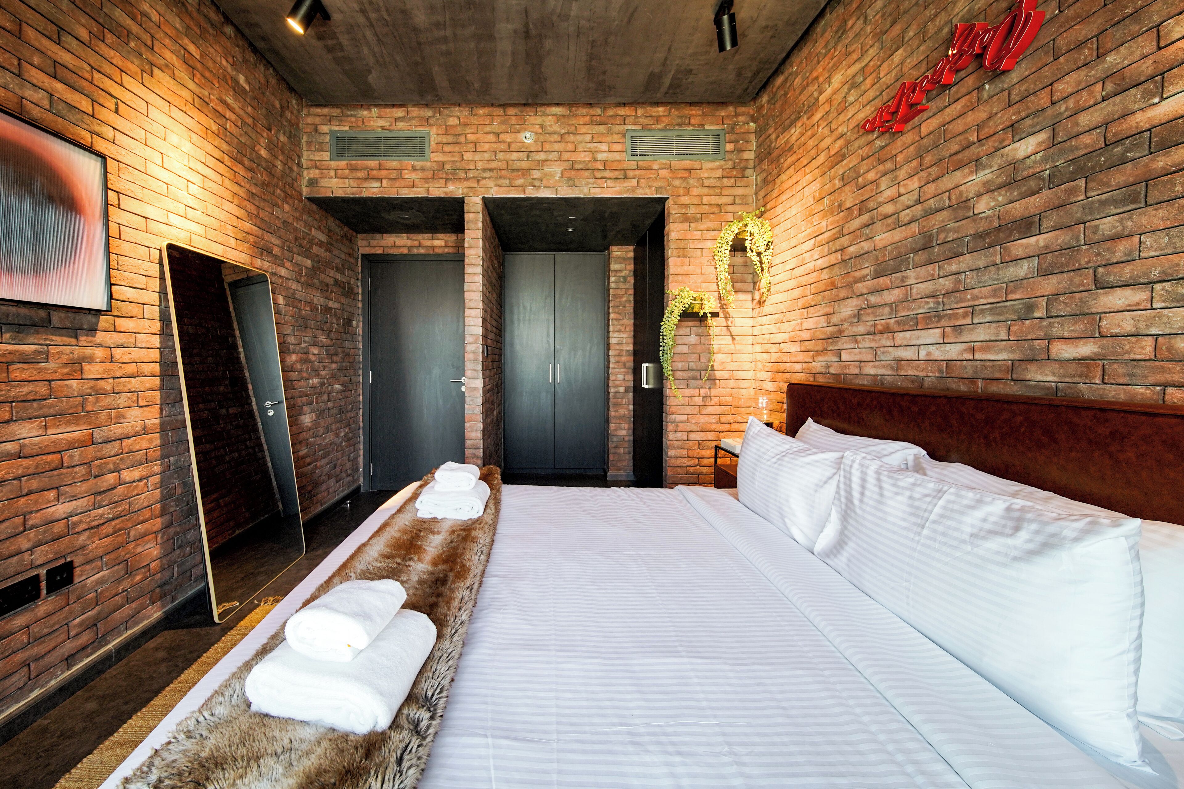 Habitació d'hotel moderna amb paret de maó, un llit gran amb roba de llit blanca, un reposacapses de fusta i un mirall al mur esquerre.