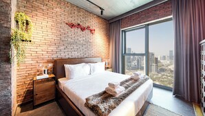 A modern hotel room with a brick wall, a large bed with white linens, a view of the city, and a small side table with a lamp.