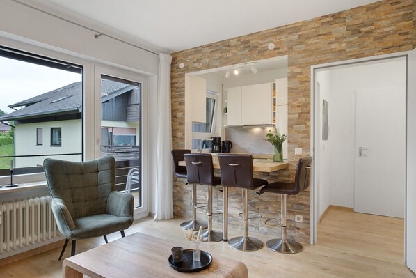 Une cuisine moderne avec un mur arrière en pierre, un coin repas avec deux tabourets de bar et une grande fenêtre donnant sur un bâtiment à l’extérieur.