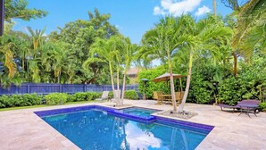 A rectangular swimming pool with a blue mosaic border, surrounded by a patio area with chairs and a table.