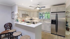 A modern kitchen with white cabinets, a stainless steel refrigerator, a built-in oven, and a ceiling fan.