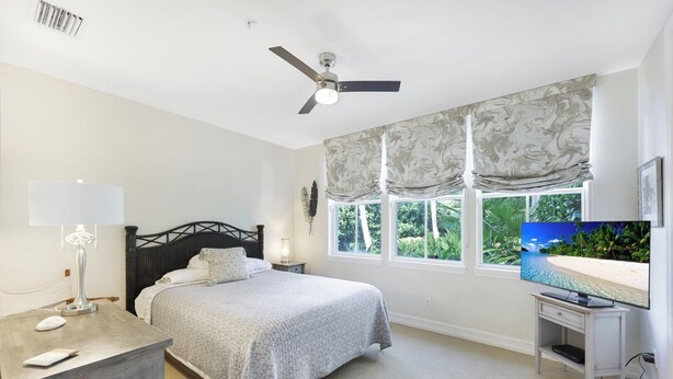 A bedroom with a bed, bedside lamp, TV, and a window with a view of greenery.