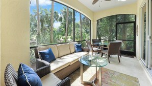 A sunlit room with a curved glass wall, a sofa with cushions, a glass table, wicker chairs, and a ceiling fan. - Bolero at Tiburon - First Floor Luxury Condo (Naples)