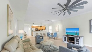 A modern living room with a beige sofa, grey armchairs, a glass coffee table, and a TV on a wooden stand.