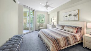 A bedroom with a large bed, a chair, a nightstand, a lamp, a ceiling fan, and a sliding glass door leading to a balcony.