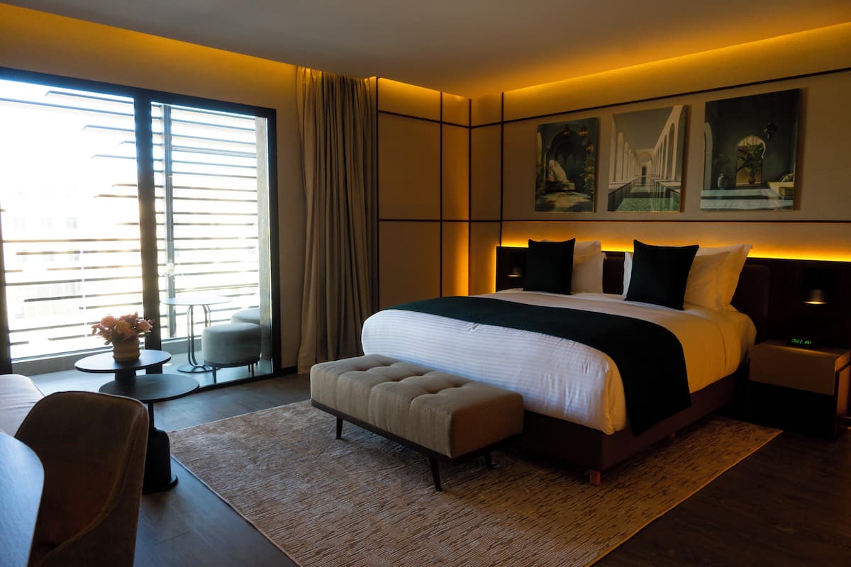 A modern hotel room with a large bed, a sitting area with a chair and a small table, and artwork on the walls.