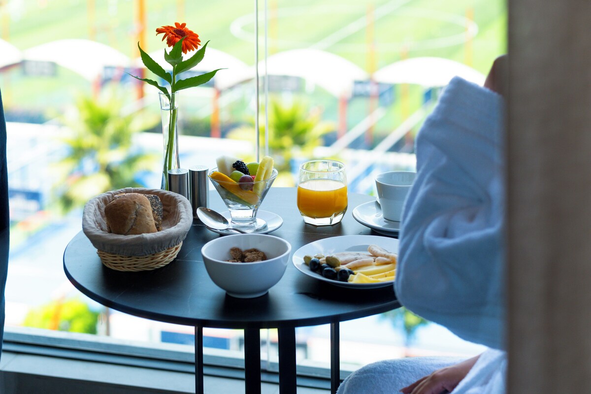 A breakfast table with a bowl of cereal, a plate of fruit, a glass of orange juice, and a vase with a flower.