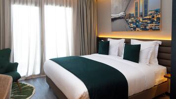 A hotel room with a large bed, a green armchair, a round rug, and a cityscape painting on the wall.