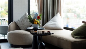 Un salon moderne avec un canapé, une petite table ronde ornée d’un vase rempli de fruits et de fleurs, et une vue sur l’extérieur grâce à de grandes fenêtres.