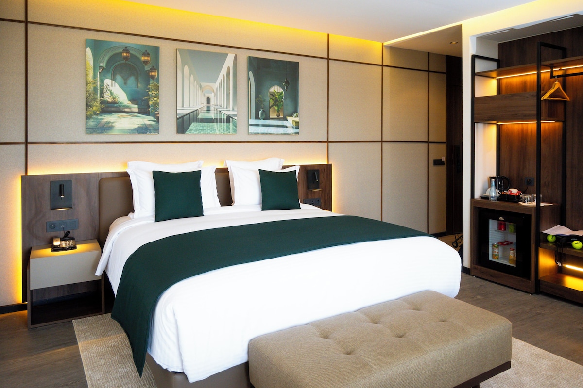 A modern hotel room with a large bed, a nightstand, a bench, and a minibar.