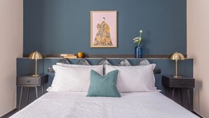Una cama bien hecha con ropa blanca y un cojín turquesa, flanqueada por dos mesitas de noche con lámparas, frente a una pared azul oscuro con una obra enmarcada encima.