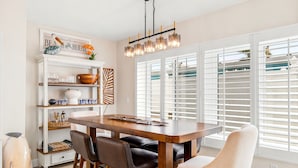 A dining area with a wooden table, chairs, a chandelier, and a window with blinds.