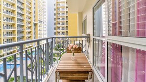 A balcony with a wooden table and a vase of flowers, overlooking a swimming pool and other buildings.