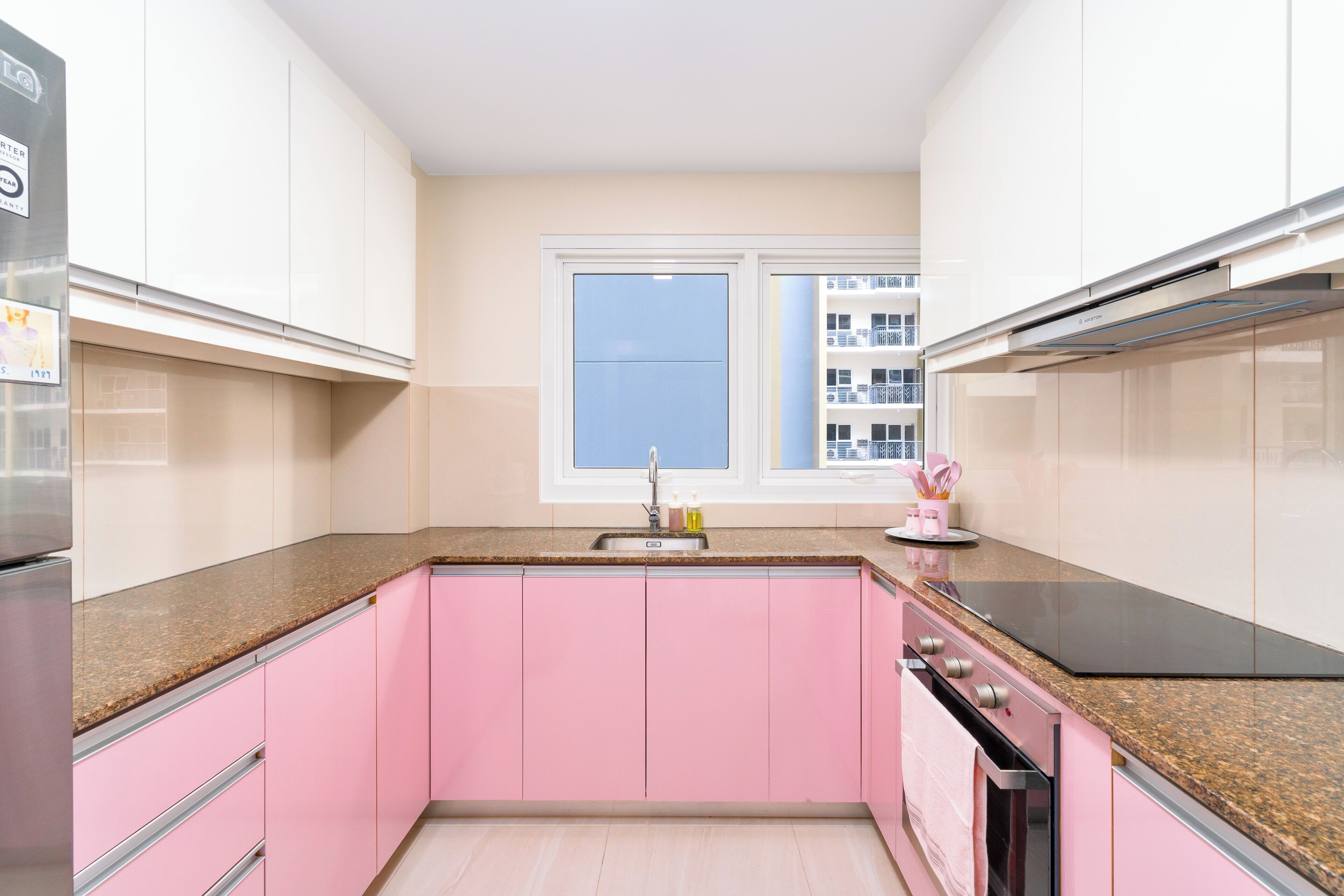 A kitchen with pink cabinets, a stainless steel refrigerator, a black stove, and a window with a view of buildings.