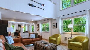 A spacious living room with a high ceiling, large windows, a brown leather sofa, a green armchair, and a wicker coffee table.