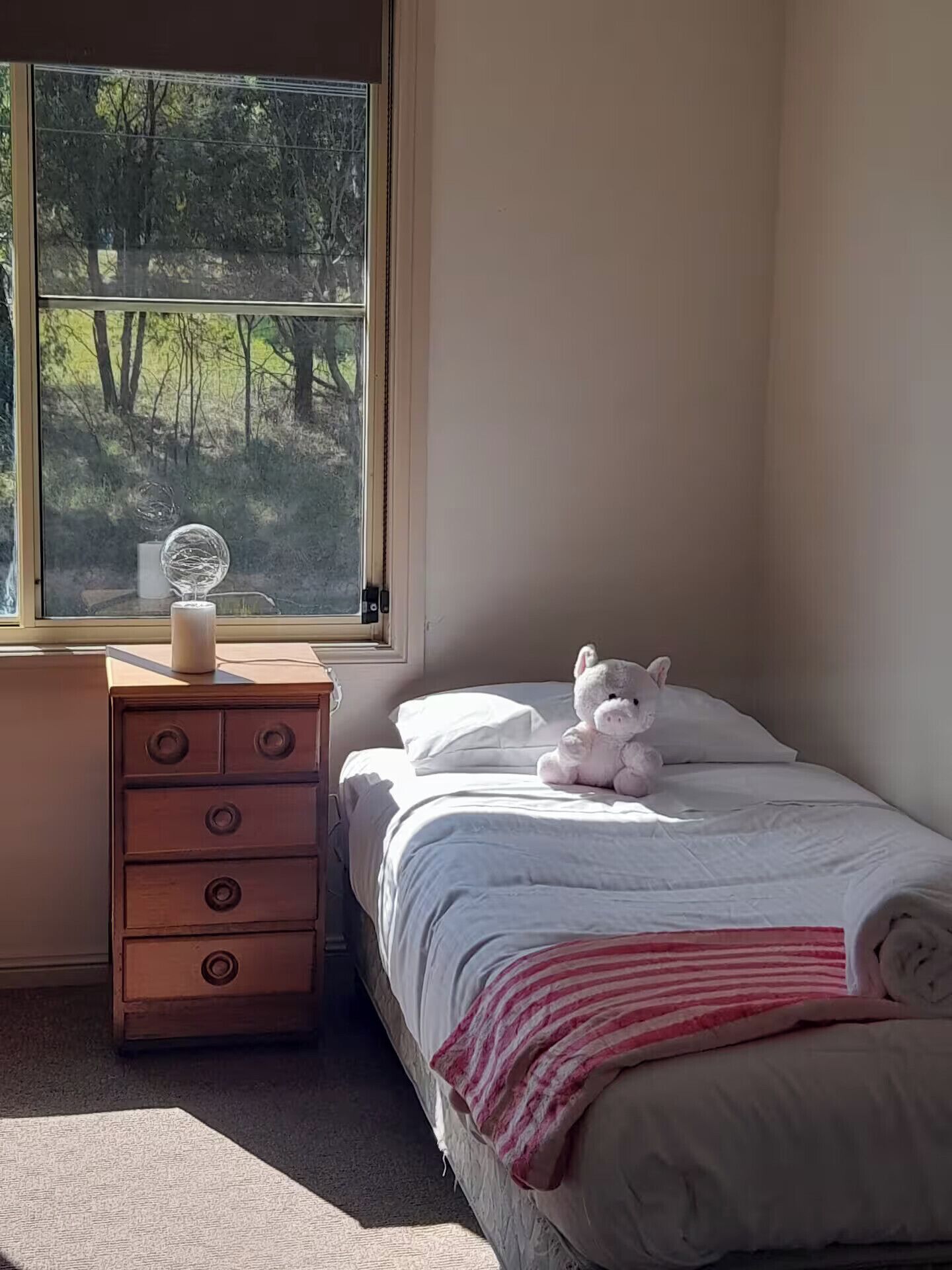 A bedroom with a bed, a dresser, a window with a view of trees, and a stuffed animal on the bed.