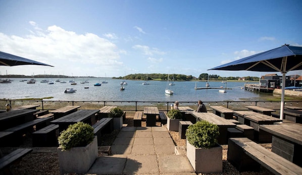 Une terrasse au bord de l’eau, avec des bancs et des tables en bois, donnant sur un lac paisible où flottent plusieurs bateaux.