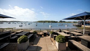 A waterfront patio with wooden benches and tables, overlooking a calm lake with several boats. - Homely Cottage for 6 Near Chichester Goodwood (Chichester)
