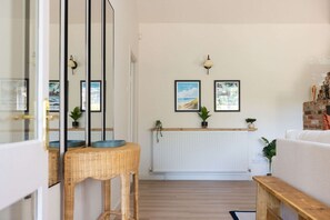 A modern living room with a white radiator, two framed pictures on the wall, a wicker side table, and a wooden bench. - Homely Cottage for 6 Near Chichester Goodwood (Chichester)