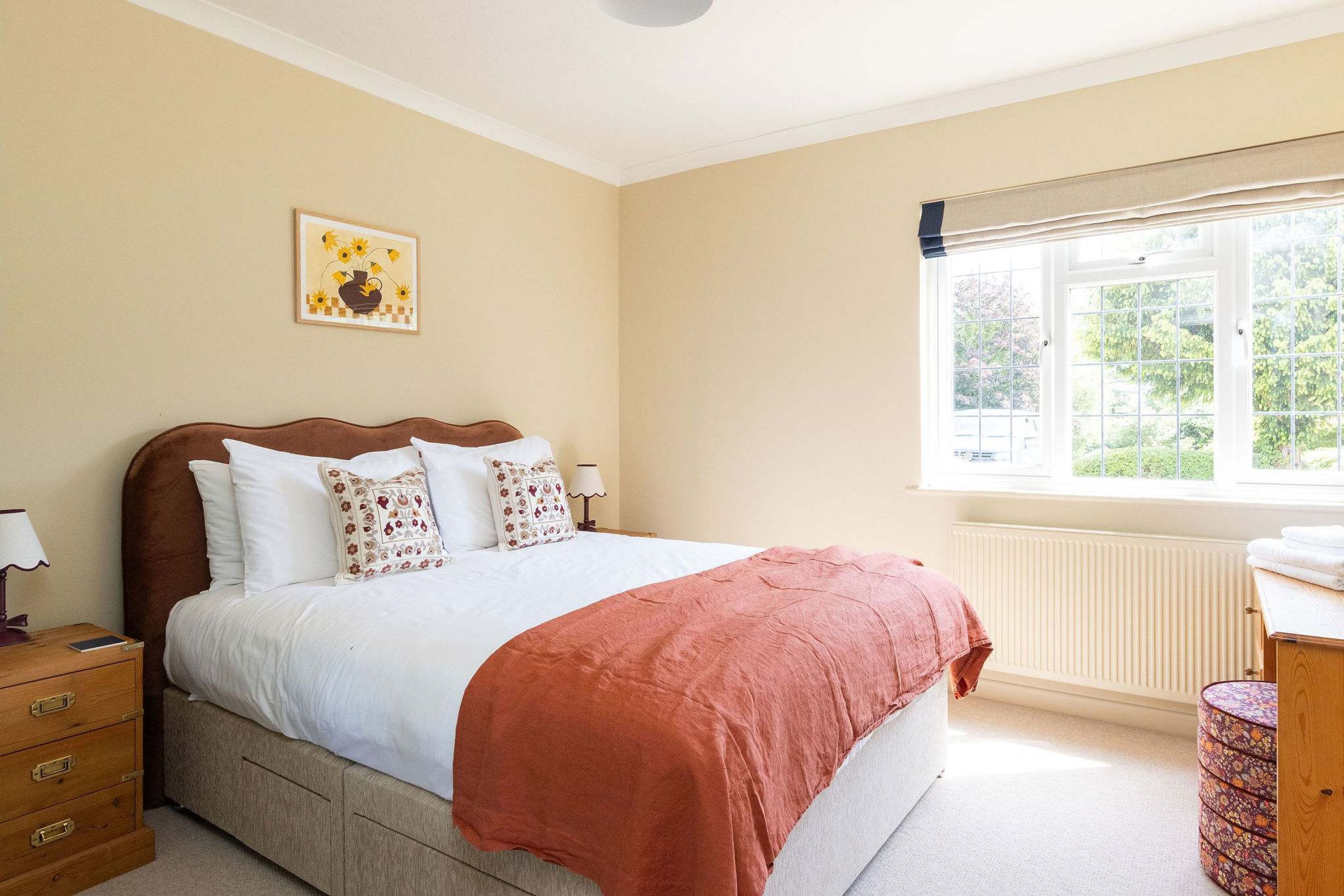 A bedroom with a bed, a nightstand, a dresser, a window with a view of greenery, and a wall-mounted picture.